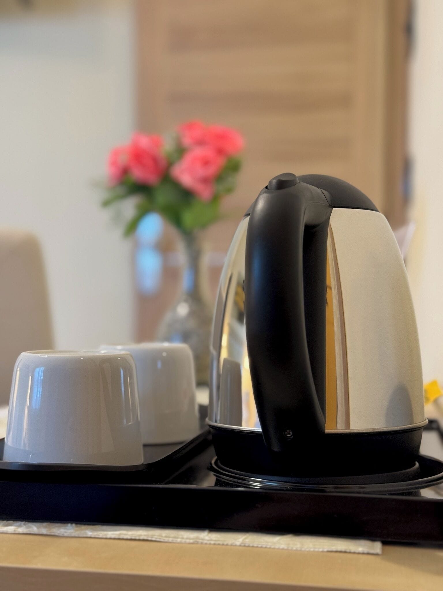 Una cafetera con dos tazas sobre una bandeja, colocada en una mesa con un florero de flores rosas al fondo.