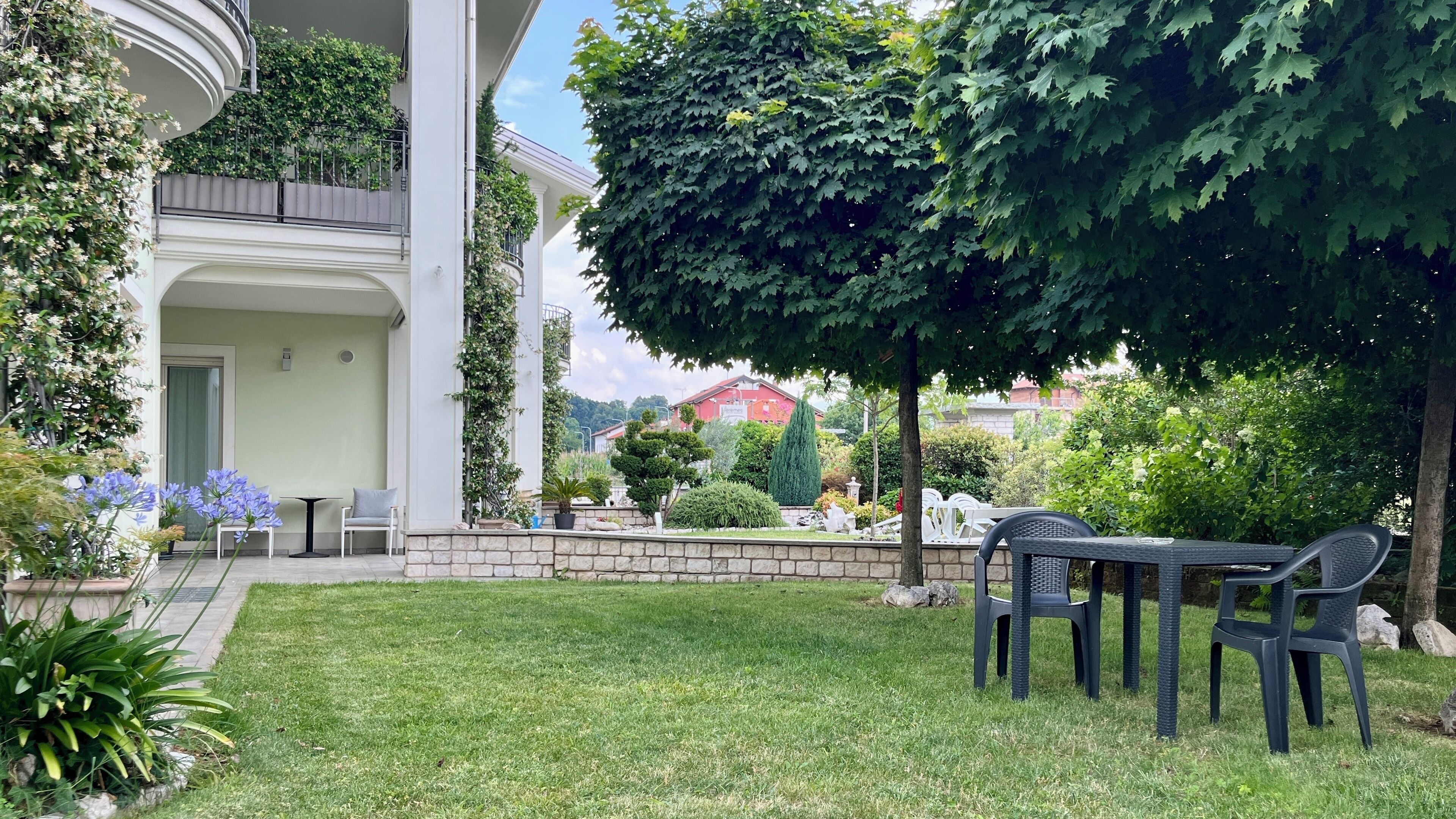 Ein gepflegter Garten mit einer Terrasse, Sitzgelegenheiten im Freien und Blick auf ein rotes Gebäude im Hintergrund.