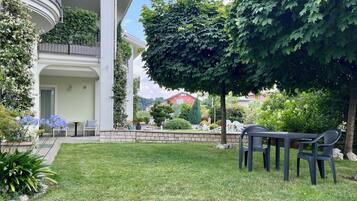 Ein gepflegter Garten mit einer Terrasse, Sitzgelegenheiten im Freien und Blick auf ein rotes Gebäude im Hintergrund.