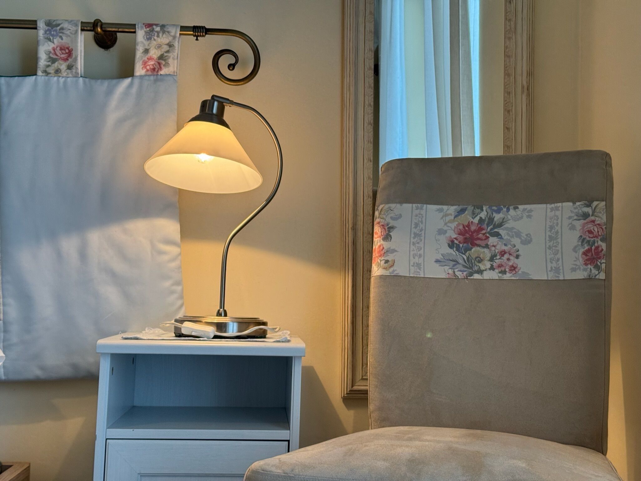 A room with a nightstand, a lamp, a chair, and a window with curtains.