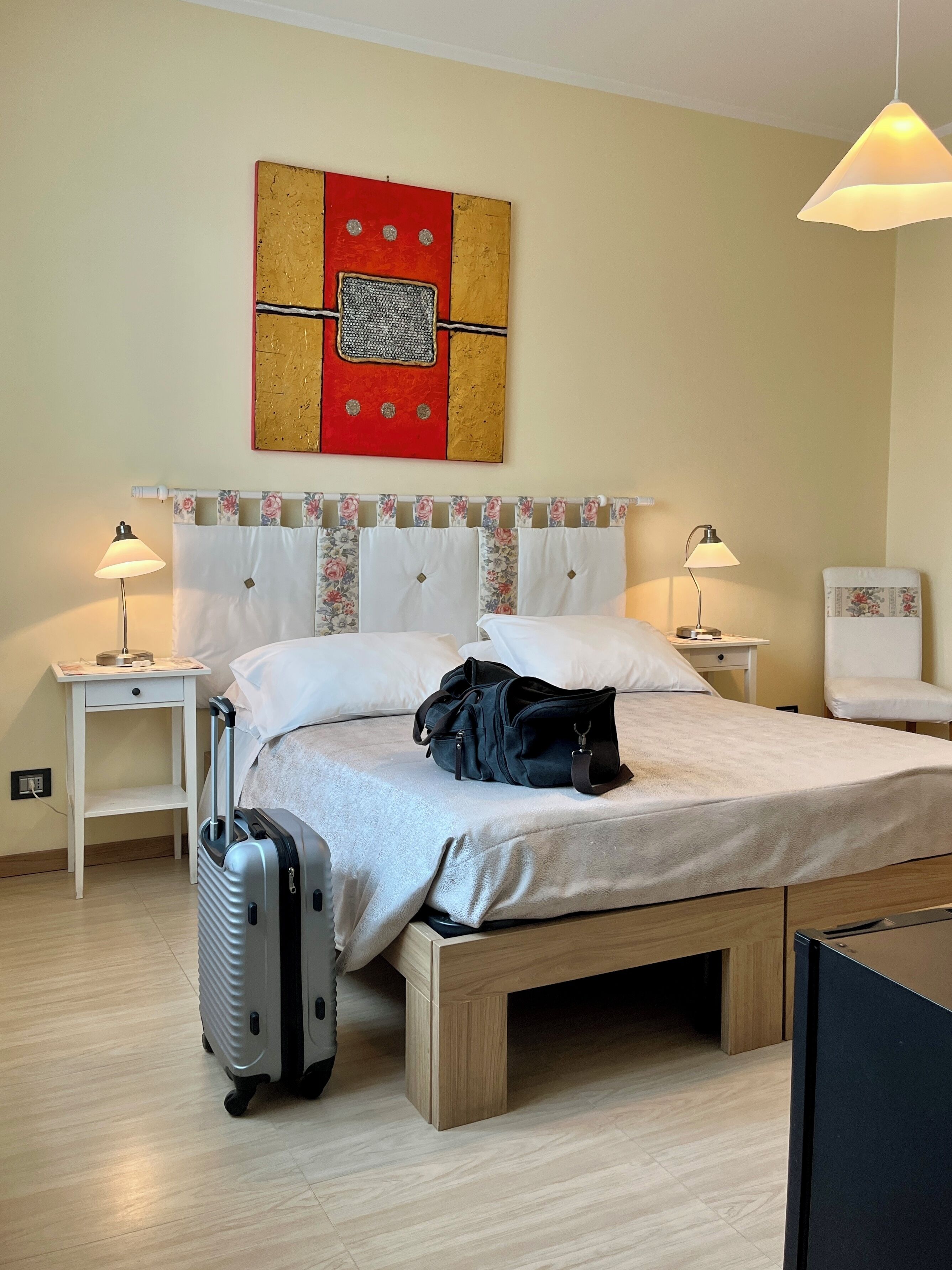 Et hotelværelse med seng, kuffert, lampe og et kunstværk på væggen.