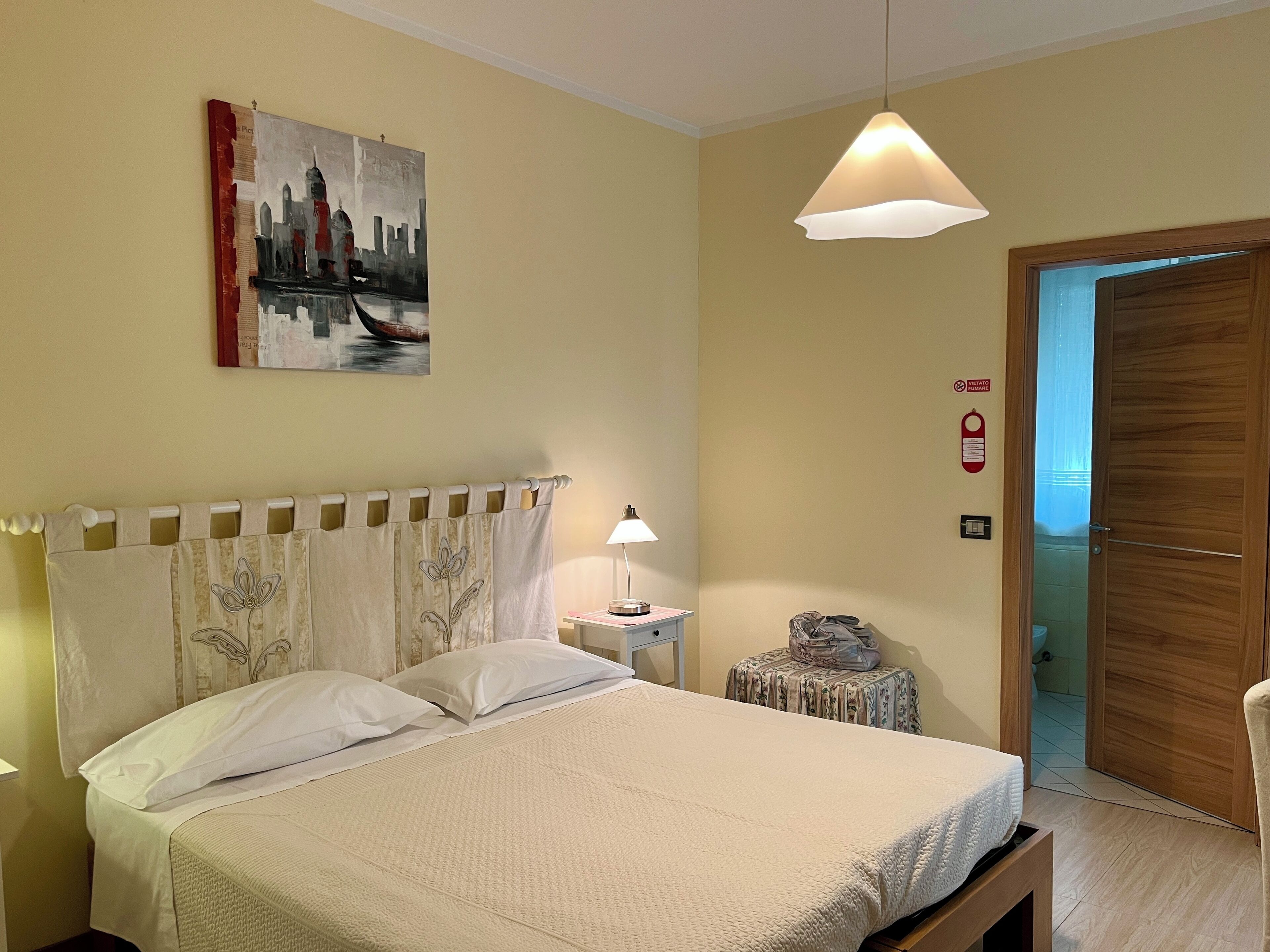 A hotel room with a bed, a nightstand, a lamp, a painting, a door, and a fire alarm.