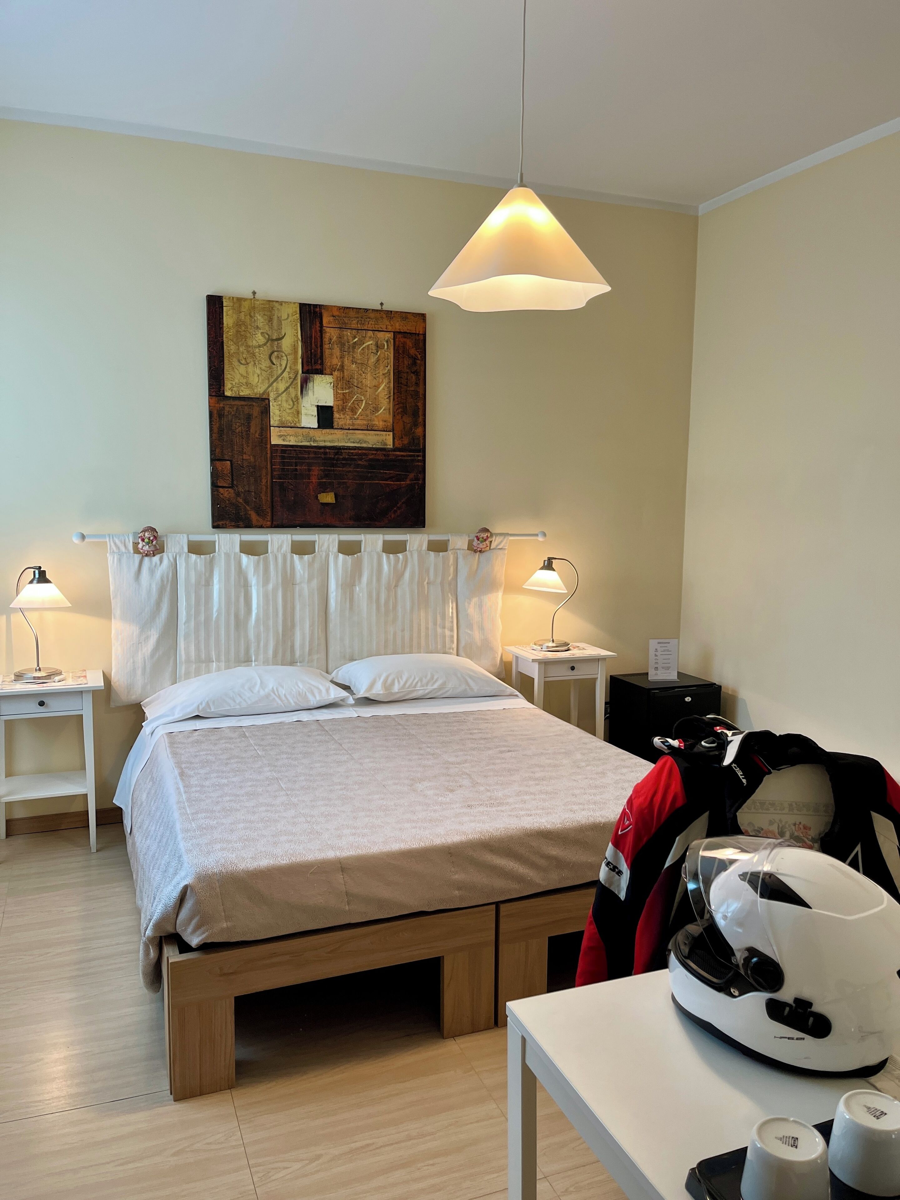 A neatly arranged hotel room with a bed, bedside lamps, a wall art piece, and a table with a suitcase and a helmet.