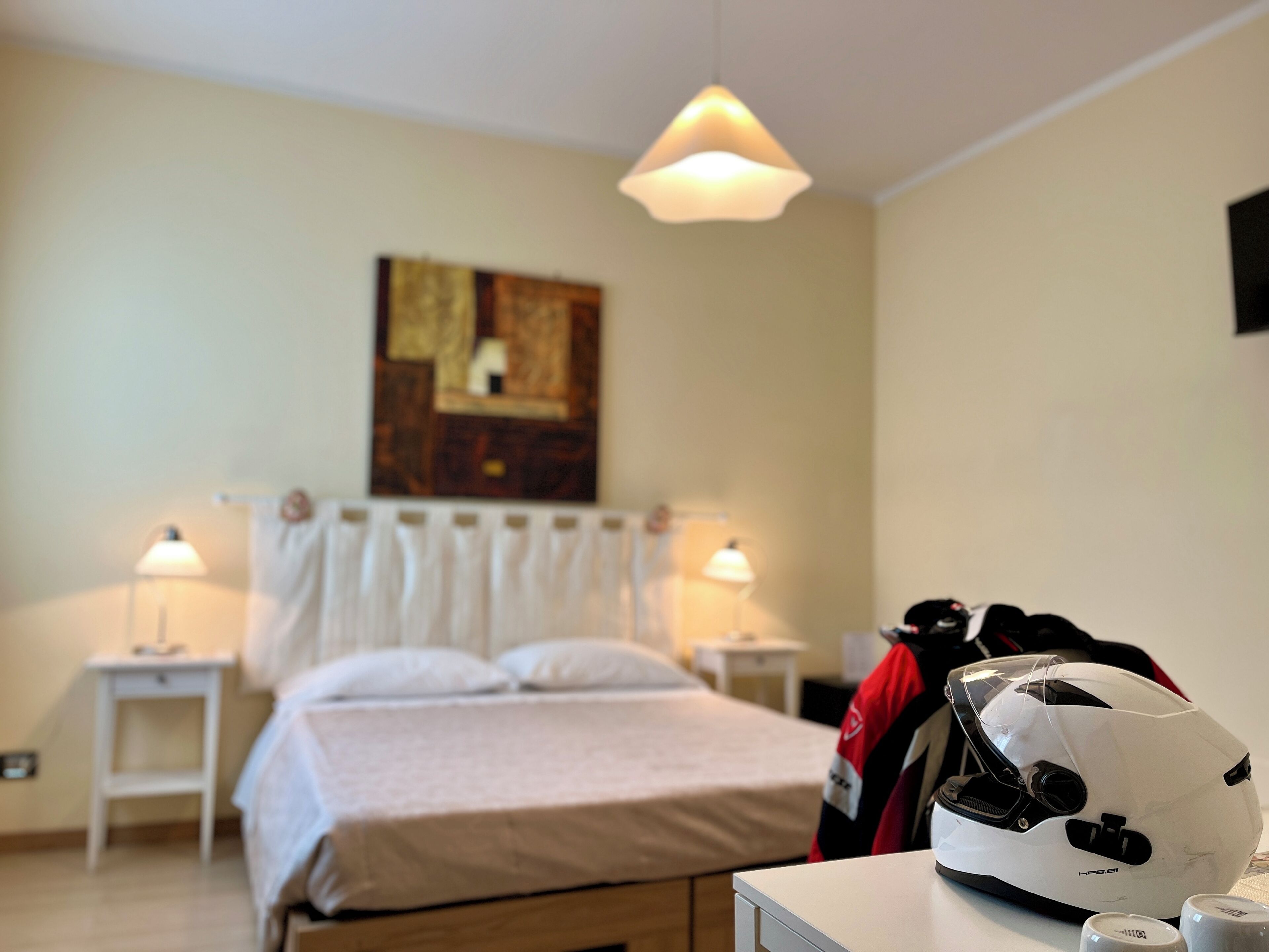 Habitación de hotel con cama, lámparas de noche, un cuadro en la pared y un casco sobre la mesa.