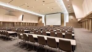 Un salón de conferencias con filas de mesas y sillas, una pantalla grande y un proyector.