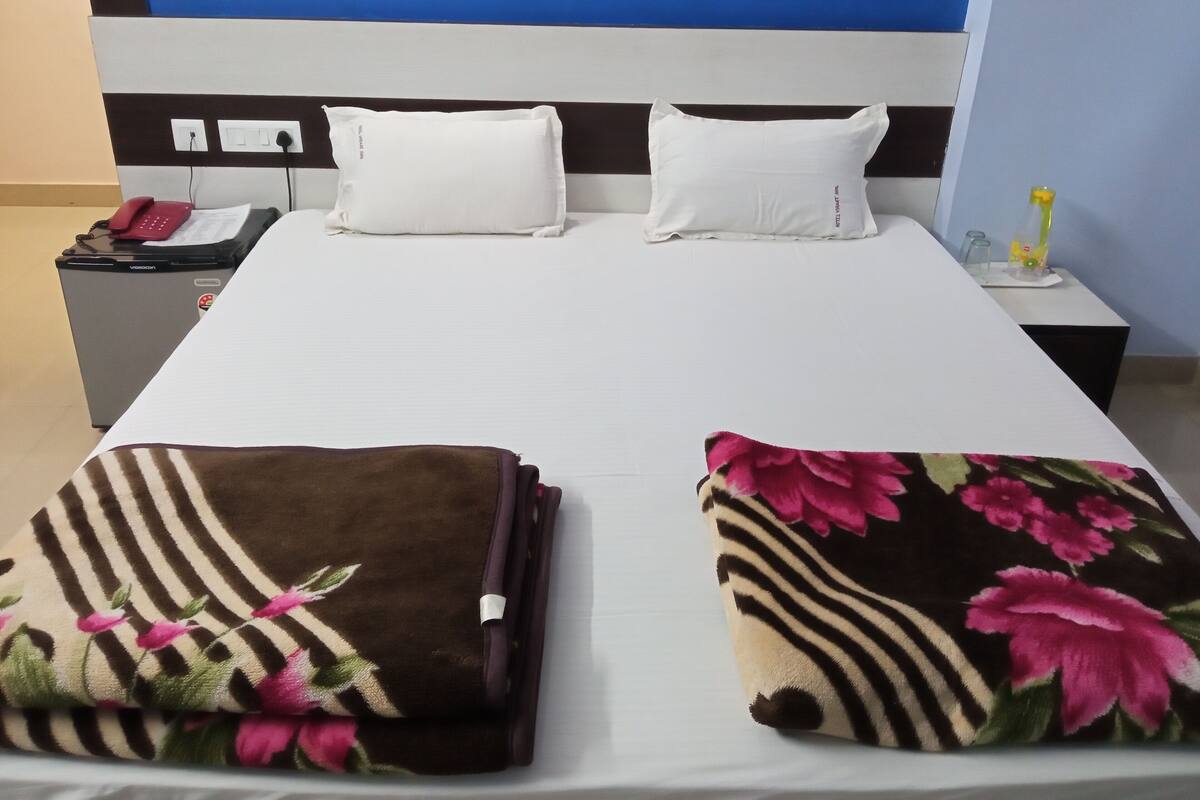 Ein Hotelzimmer mit einem Bett, zwei Kissen, einem Nachttisch mit Telefon, einer Lampe und einem kleinen Tisch mit einer Flasche.