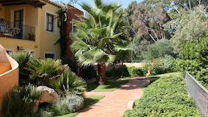 Ein gelbes Gebäude mit Balkon, umgeben von einem Garten mit Palmen und Sträuchern.