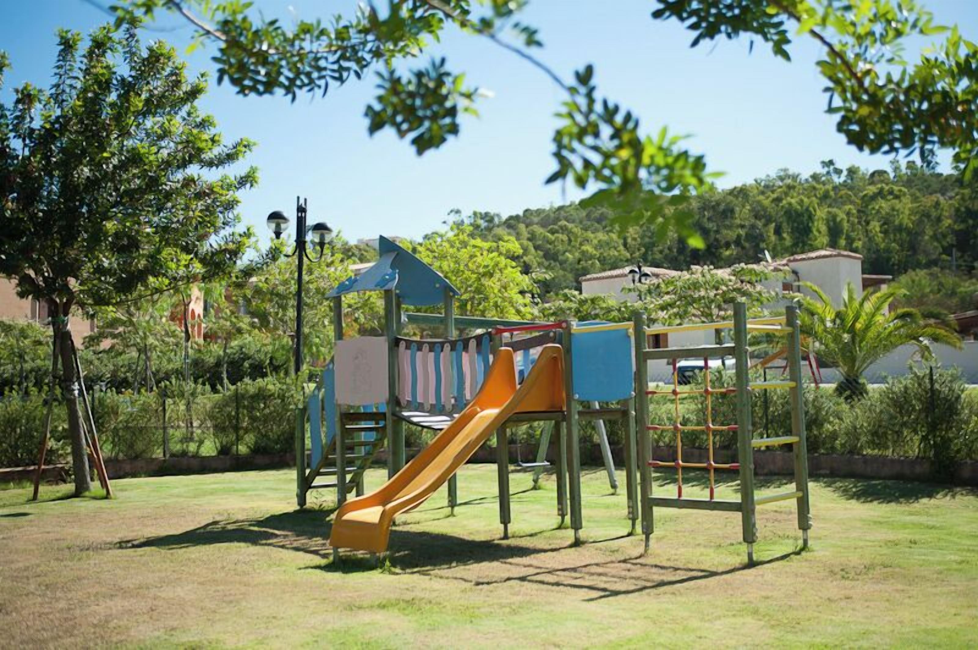A playground with a slide and climbing structures, surrounded by trees and buildings in the background.