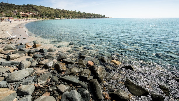 Ein felsiger Strand mit klarem Wasser, eine entfernte Küstenlinie mit Gebäuden und einem wolkenlosen Himmel.