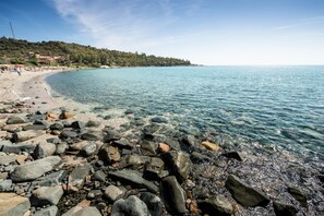 Une plage rocheuse aux eaux cristallines, une côte lointaine parsemée de bâtiments et un ciel dégagé.