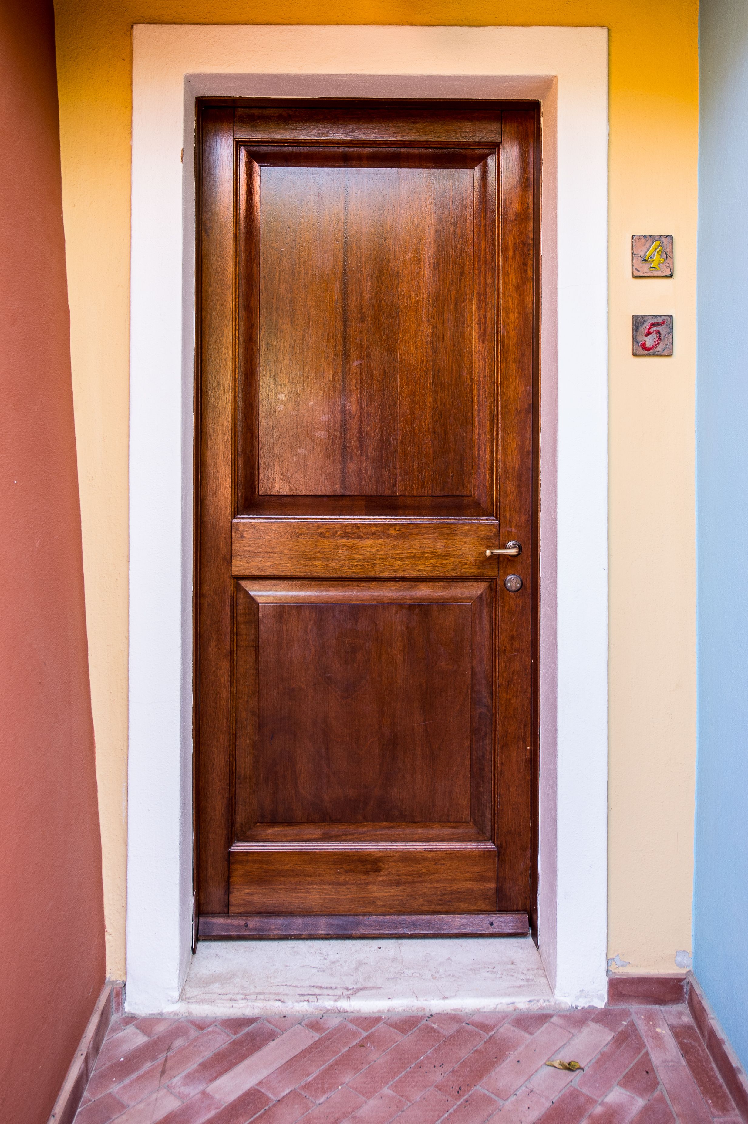 A wooden door with a brass handle, set in a hallway with a tiled floor and a wall with a number plaque.