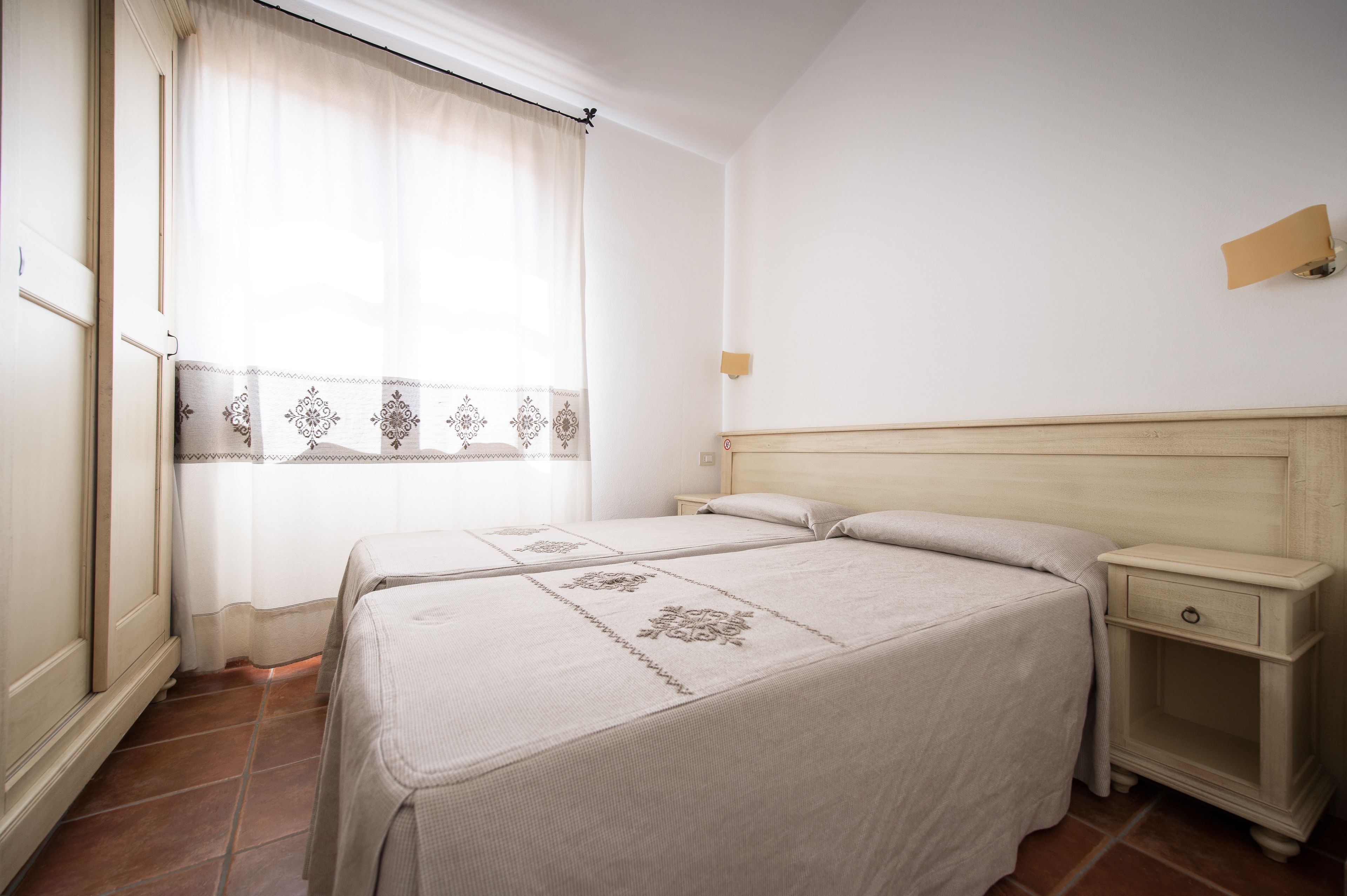 Un dormitorio con dos camas, una mesita de noche, una lámpara de pared y una ventana con cortinas.