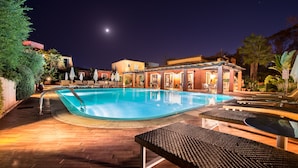 Ein Poolbereich bei Nacht mit Liegestühlen und Sonnenschirmen, umgeben von Gebäuden und Palmen.