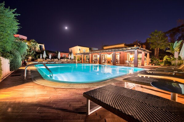 Un área de piscina nocturna con sillones de descanso y sombrillas, rodeada de edificios y palmeras.
