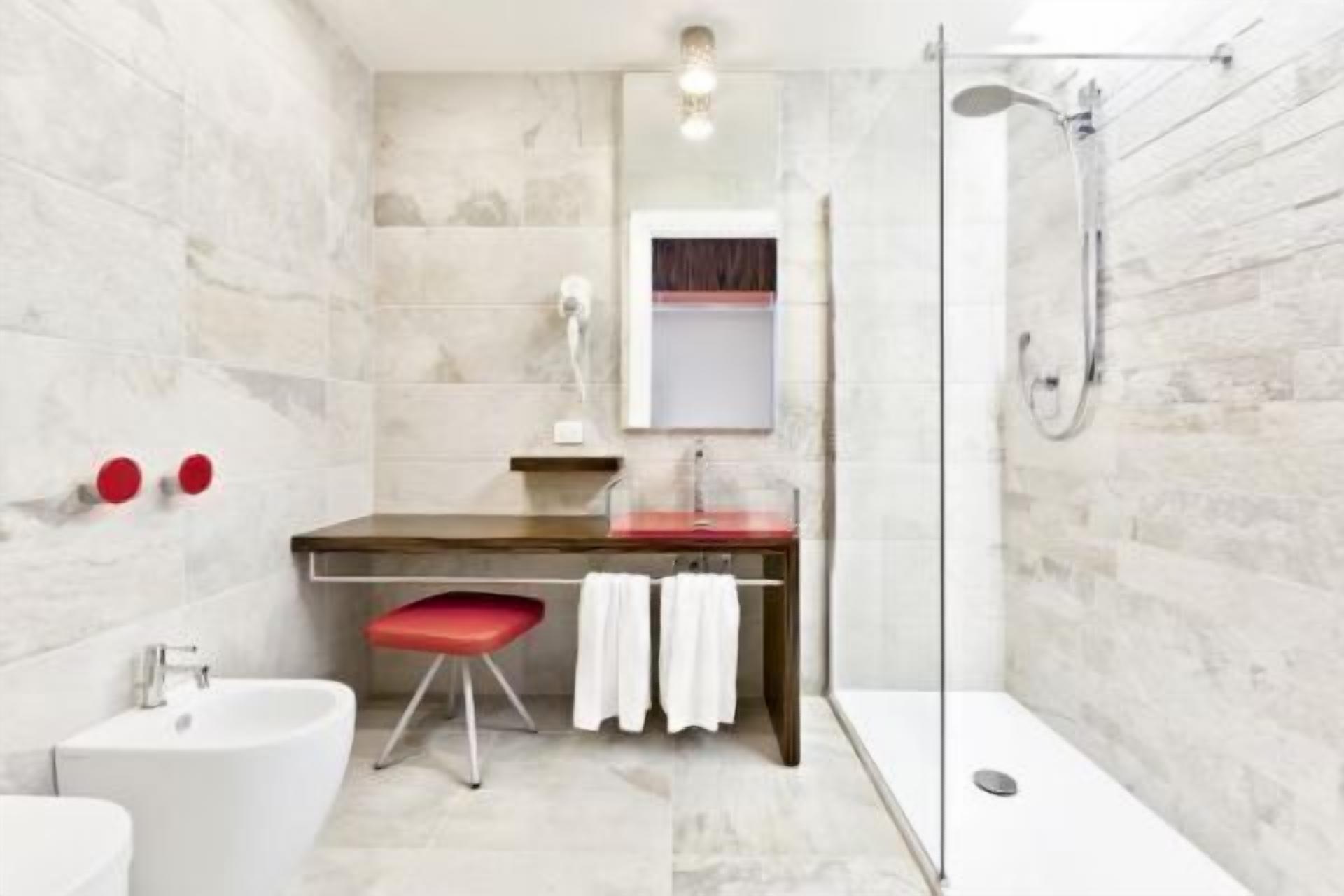 Un bagno moderno con parete in marmo, mobile lavabo in legno, uno sgabello rosso, lavandino bianco e box doccia in vetro.