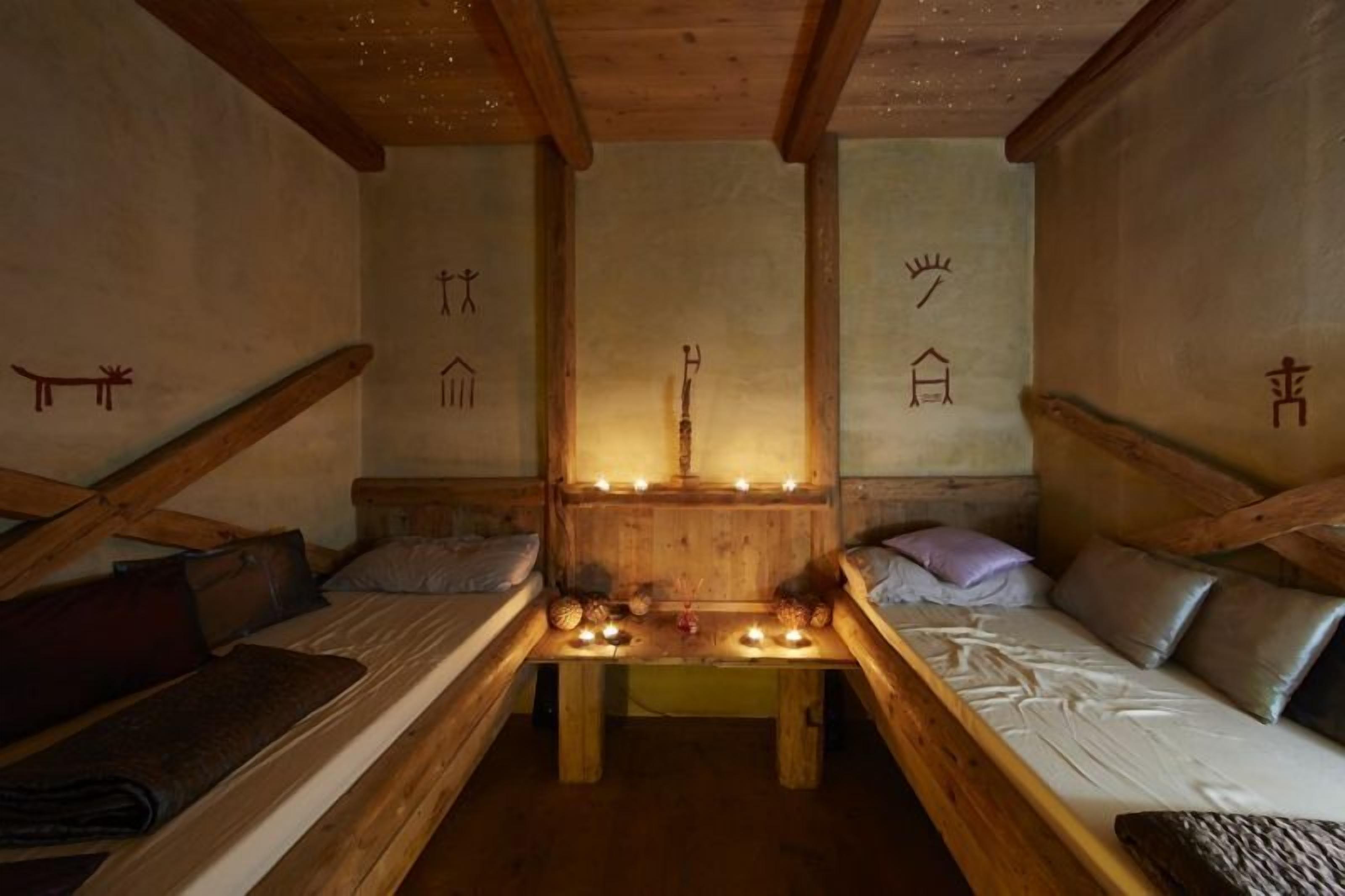 A cozy, rustic room with wooden beams, a central table with candles, and simple bed arrangements.