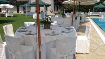 Un área de comedor al aire libre con mesas redondas dispuestas para una comida, rodeada de sillas blancas y cubierta por sombrillas.