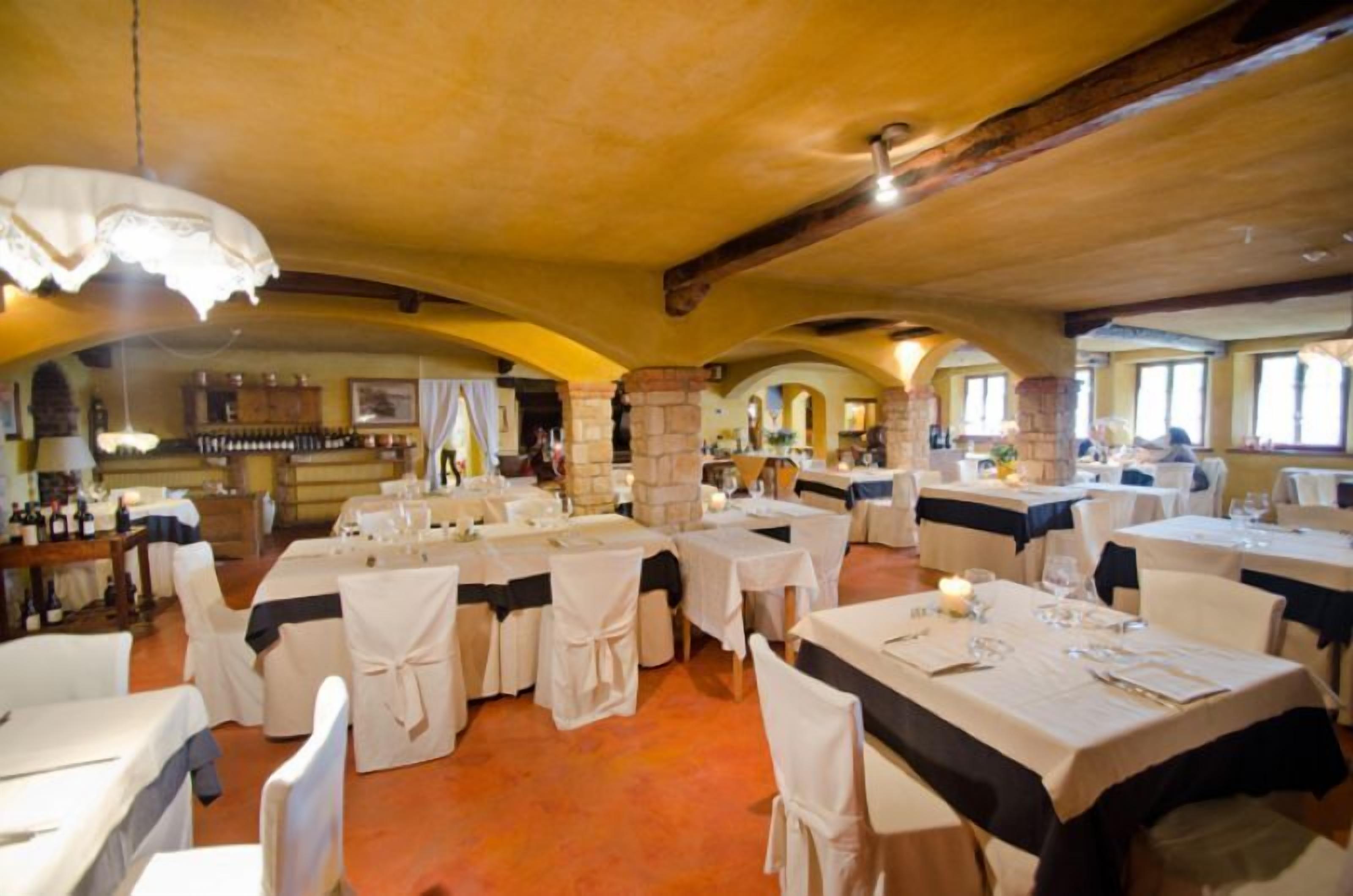 A restaurant with multiple tables and chairs, each covered with a white tablecloth and black sashes. The room has a rustic stone ceiling and walls, with a warm, orange-toned floor.
