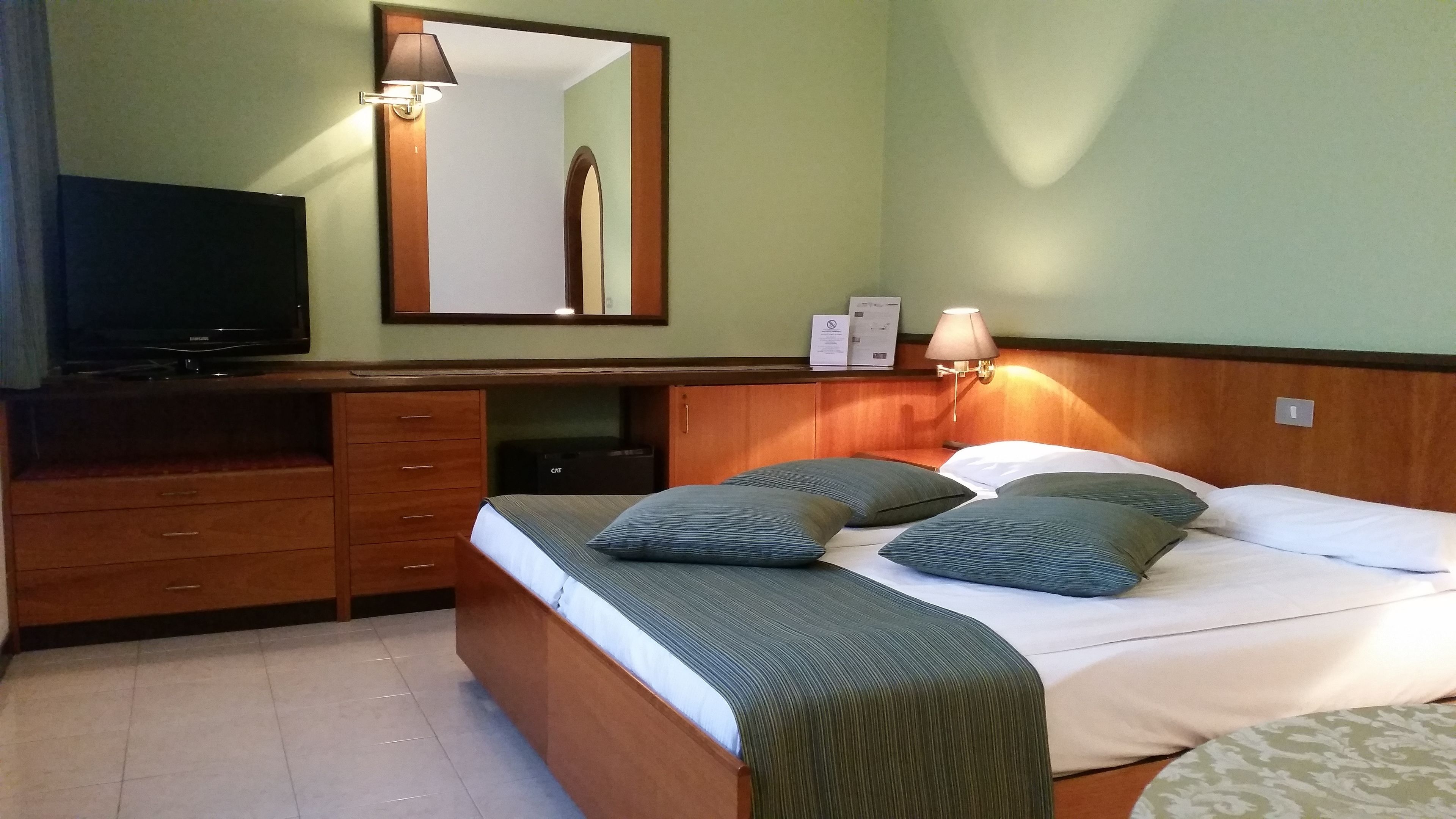 A hotel room with a bed, a TV, a desk with drawers, a mirror, and a lamp.