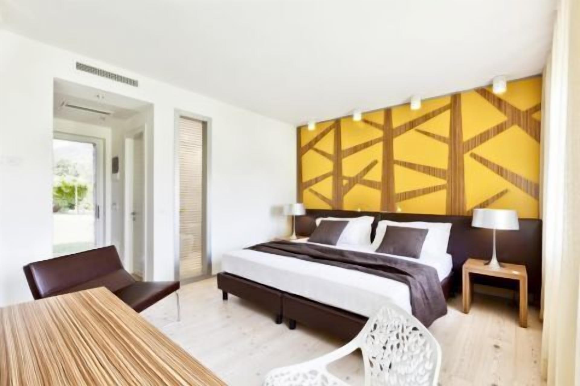Quarto de hotel moderno com uma cama grande, uma mesa de jantar e uma cadeira. Há uma janela com vista para o verde lá fora.