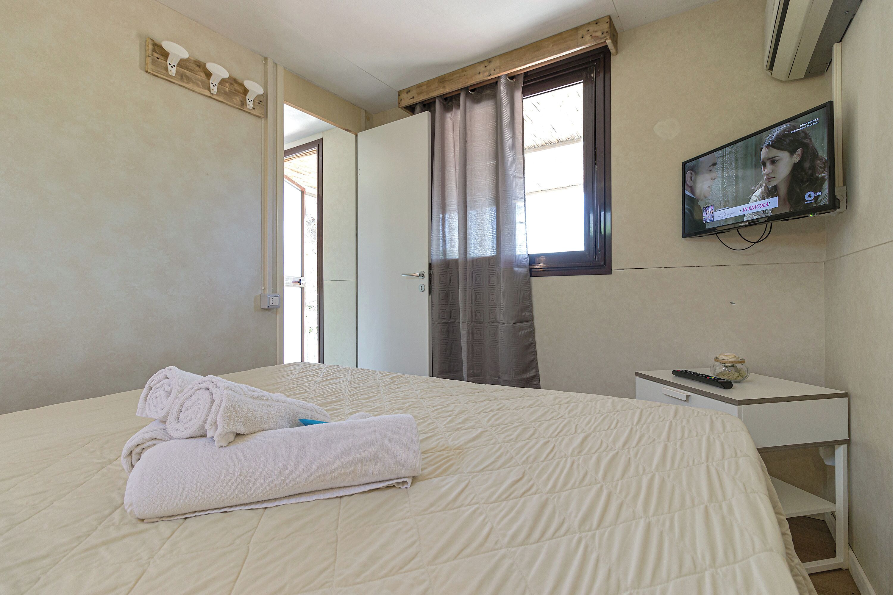 A hotel room with a bed, a TV mounted on the wall, a bedside table, and a window with curtains.