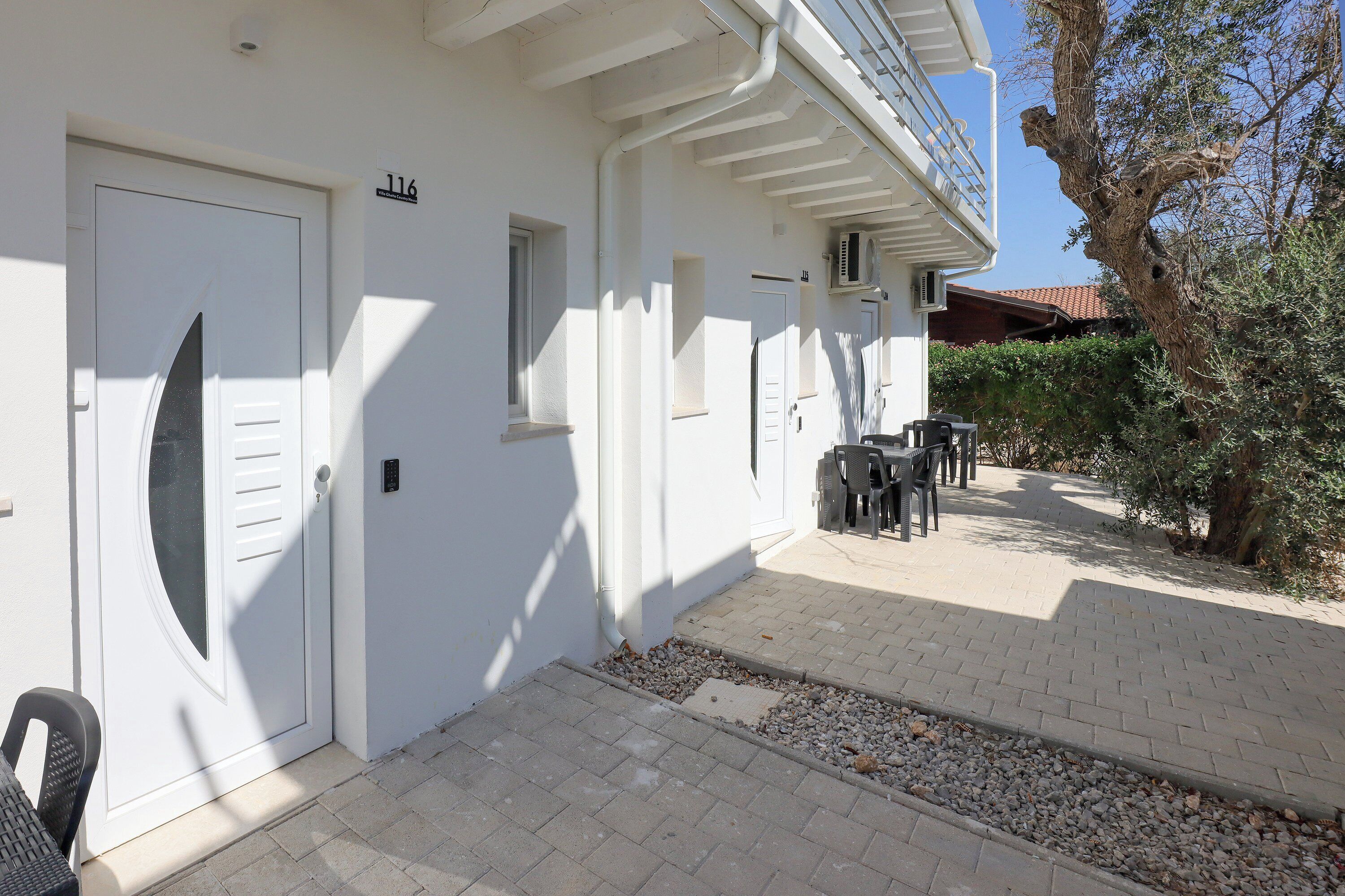 A white building with numbered doors, a paved patio, and outdoor furniture.