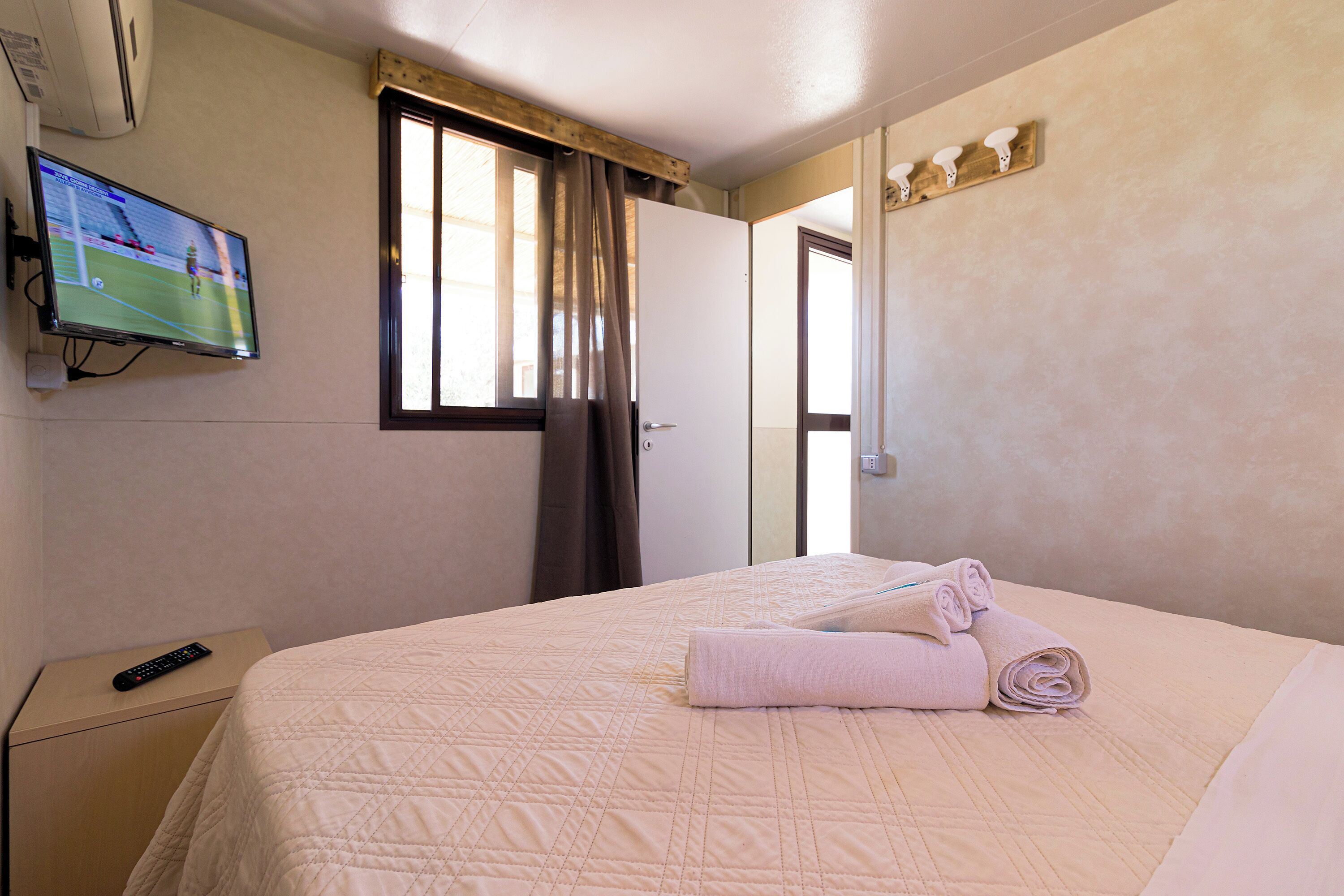 A hotel room with a bed, a TV mounted on the wall, a window with curtains, and a towel on the bed.