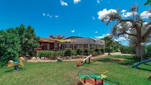 A playground with swings, a seesaw, and a climbing structure. There is a building with a red facade and a white roof in the background. - Villa Ghetta Country House (Leverano)
