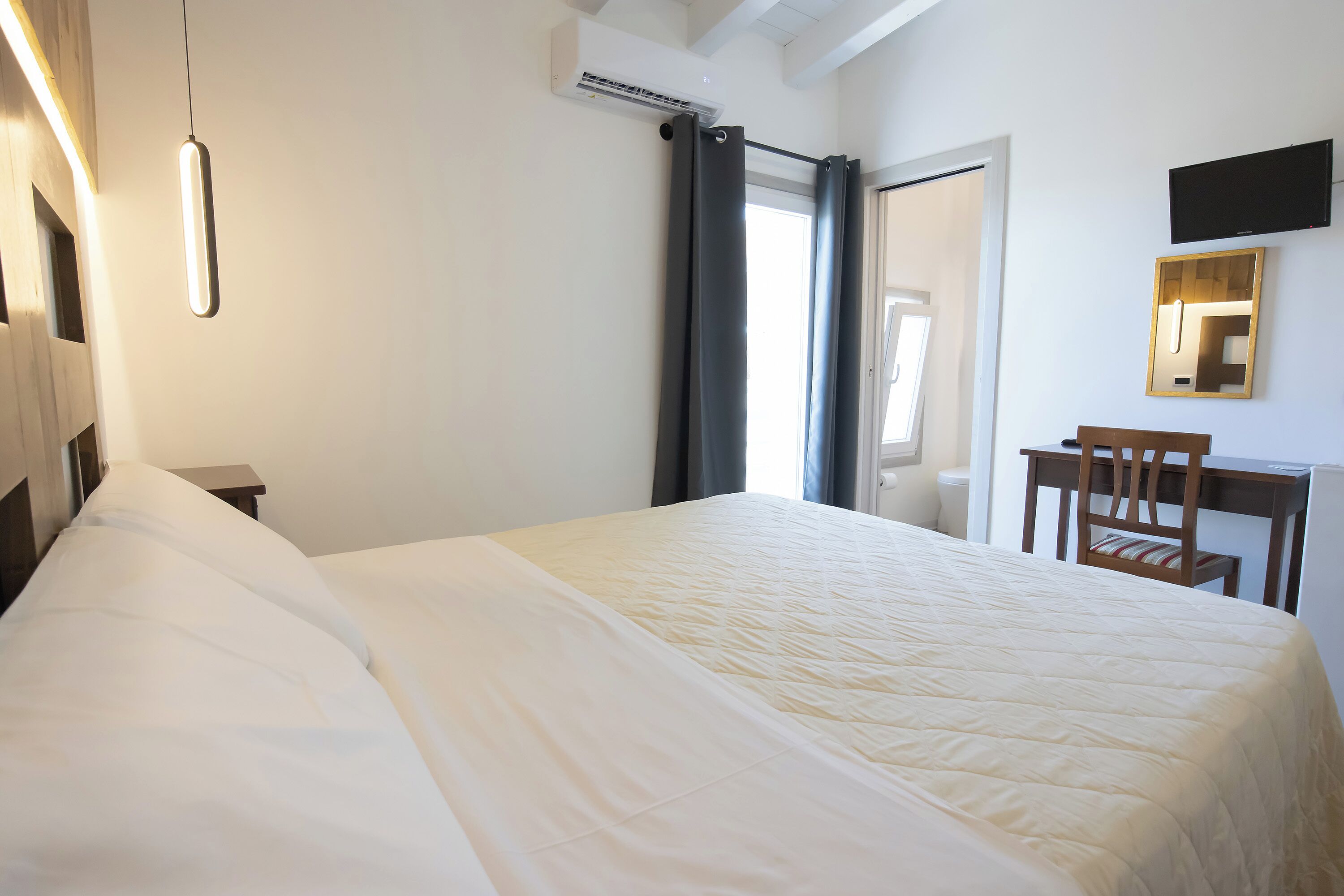 A neatly made bed with a white duvet, a bedside table, a chair, a desk, a flat-screen TV mounted on the wall, and a window with curtains.