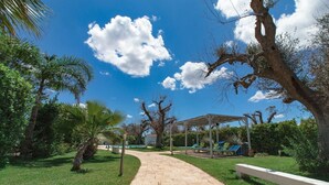 A paved pathway leading to a covered seating area with benches and chairs, surrounded by lush greenery and trees. - Villa Ghetta Country House (Leverano)