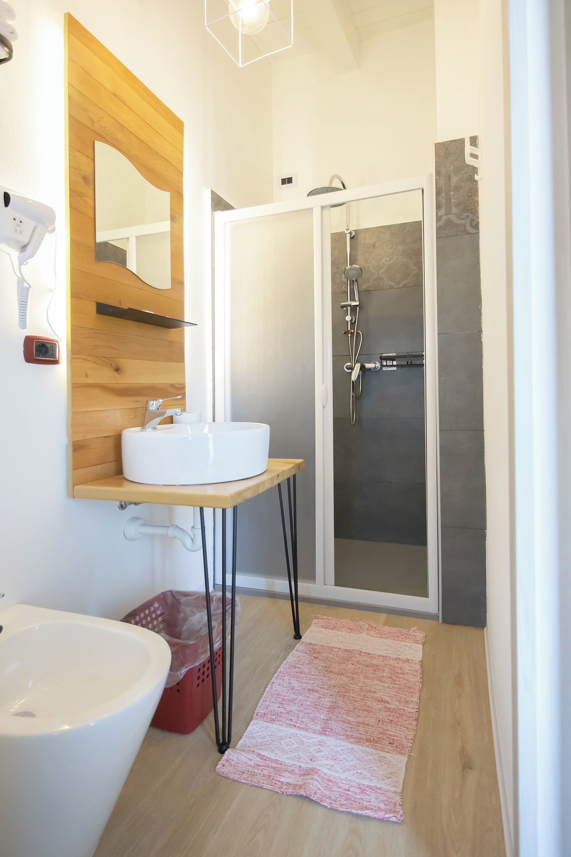 Ein Badezimmer mit einem Holzkosmetikschrank, einem weißen Waschbecken, einem Spiegel, einer Duschkabine und einem roten Wäschekorb.
