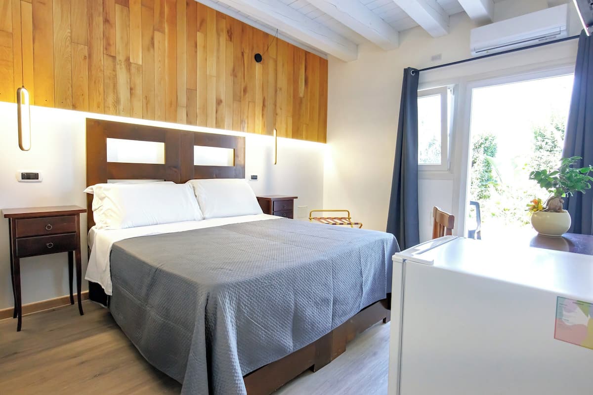 A bedroom with a wooden headboard, a bed with a grey bedspread, a bedside table, a small desk with a plant, and a window with curtains.