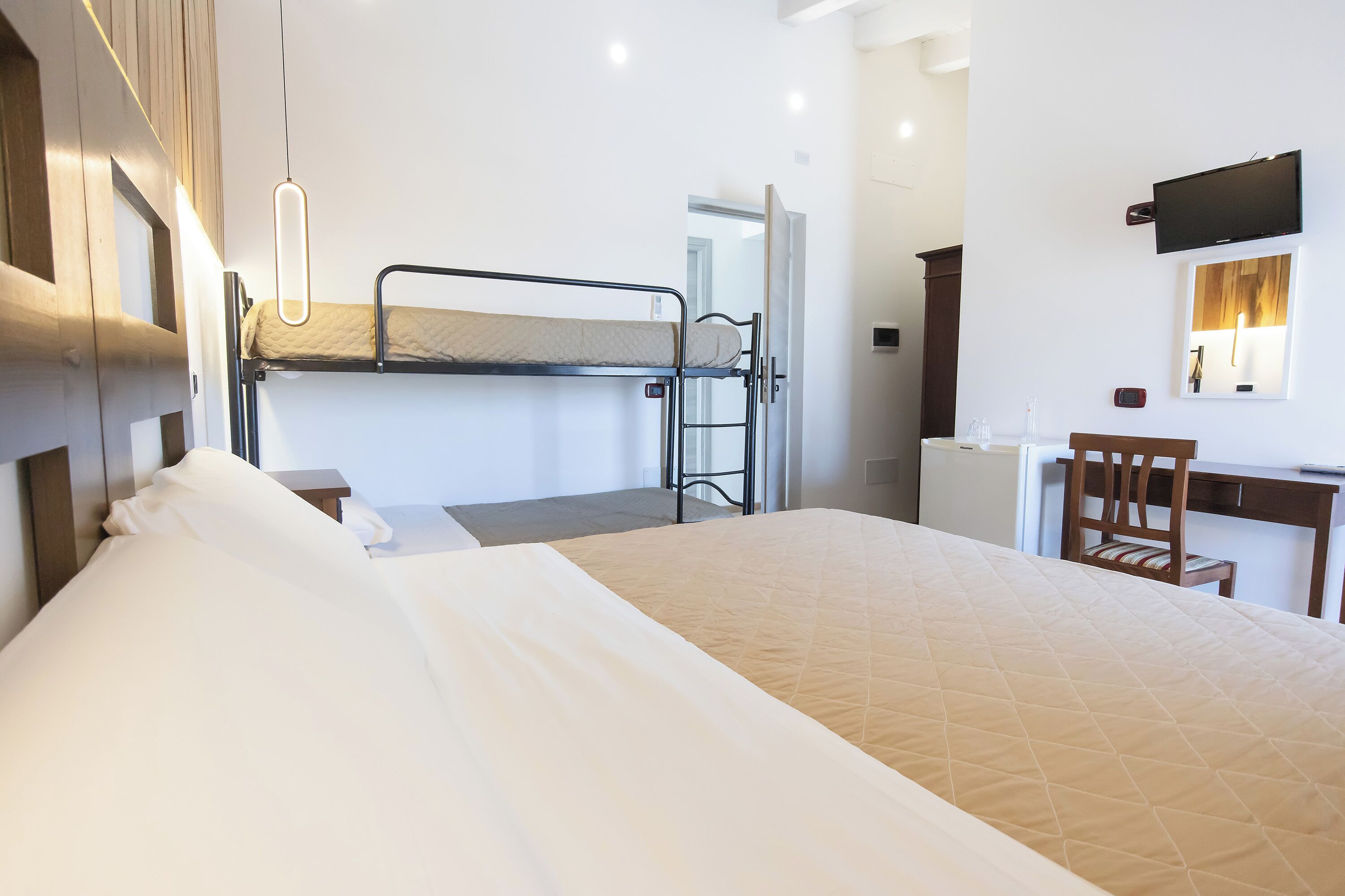 Quarto de hotel com cama de solteiro, escrivaninha, cadeira, televisão presa na parede e janela.