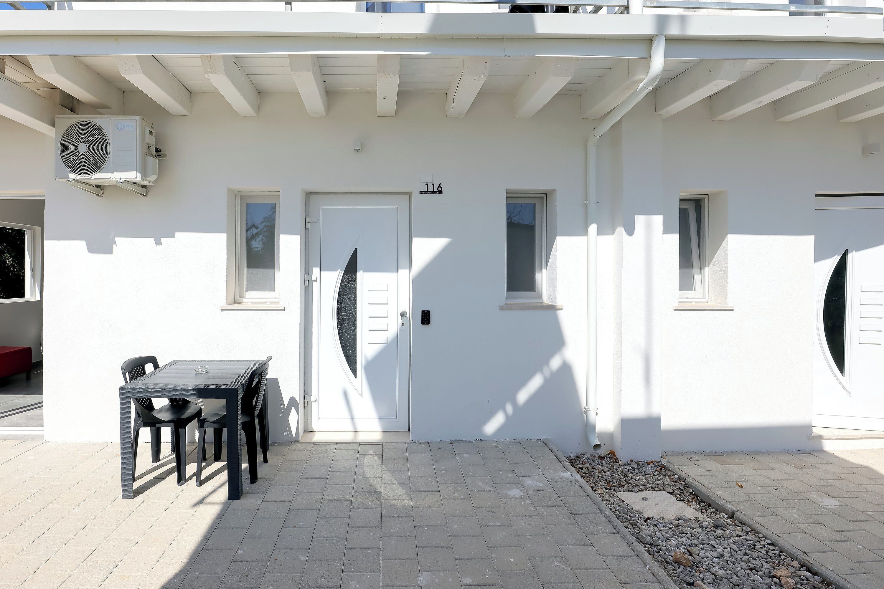 A white building with a door marked "116", a table with chairs, and an air conditioning unit mounted on the wall.