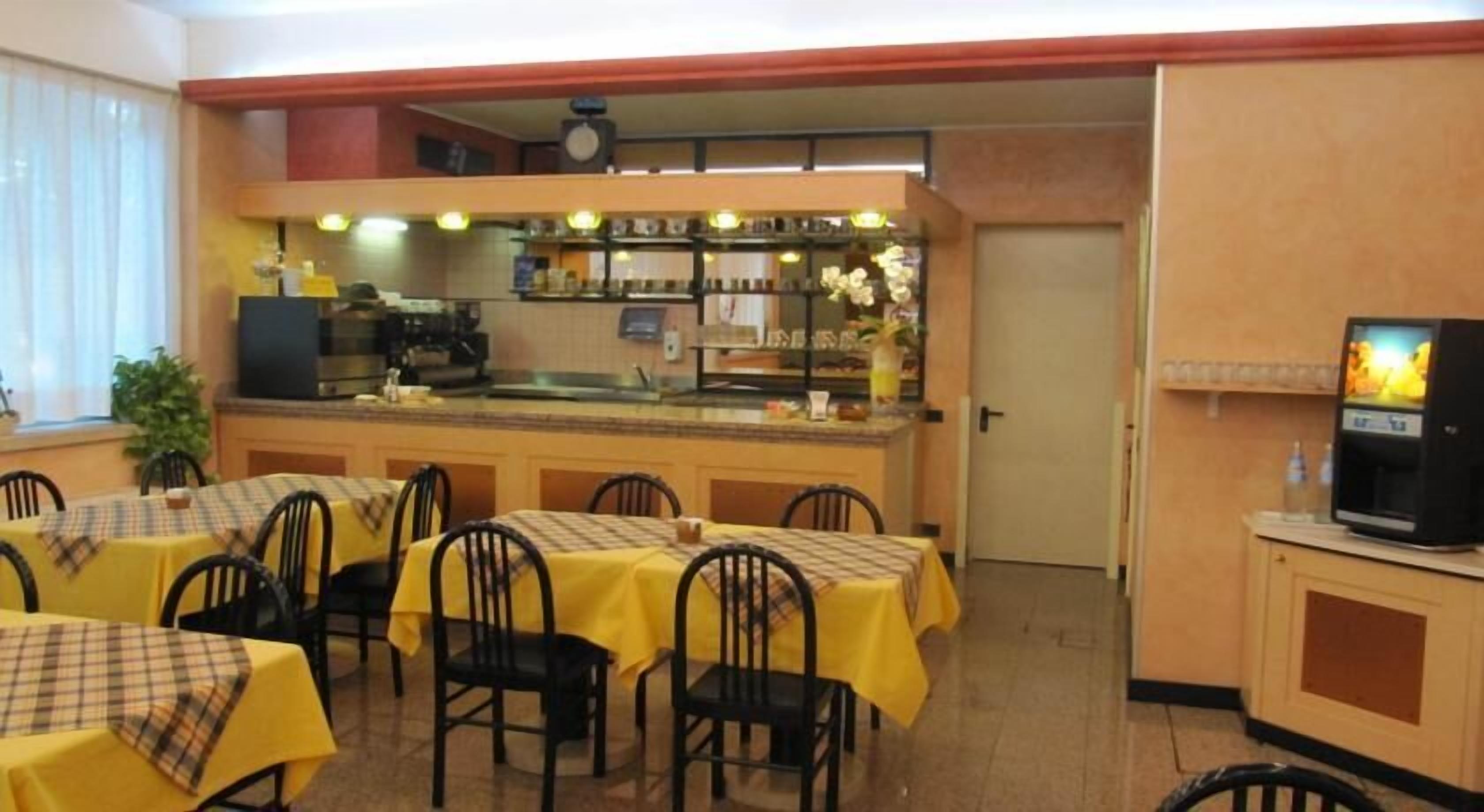 Un restaurant avec des tables recouvertes de nappes jaunes et des chaises noires. On y trouve un comptoir avec une machine à café et un écran de télévision.