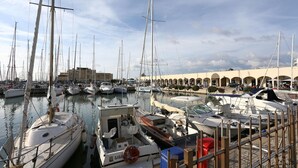Un puerto deportivo con varios veleros amarrados, un edificio con arcos al fondo y un cielo despejado.