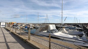 Un puerto deportivo con varias embarcaciones amarradas, un paseo marítimo pavimentado y una vista distante de un edificio colorido.