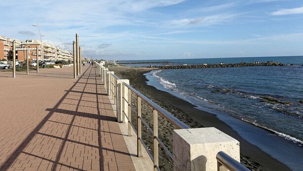 Un camino pavimentado a lo largo de la costa, con vista al mar y edificios al fondo.