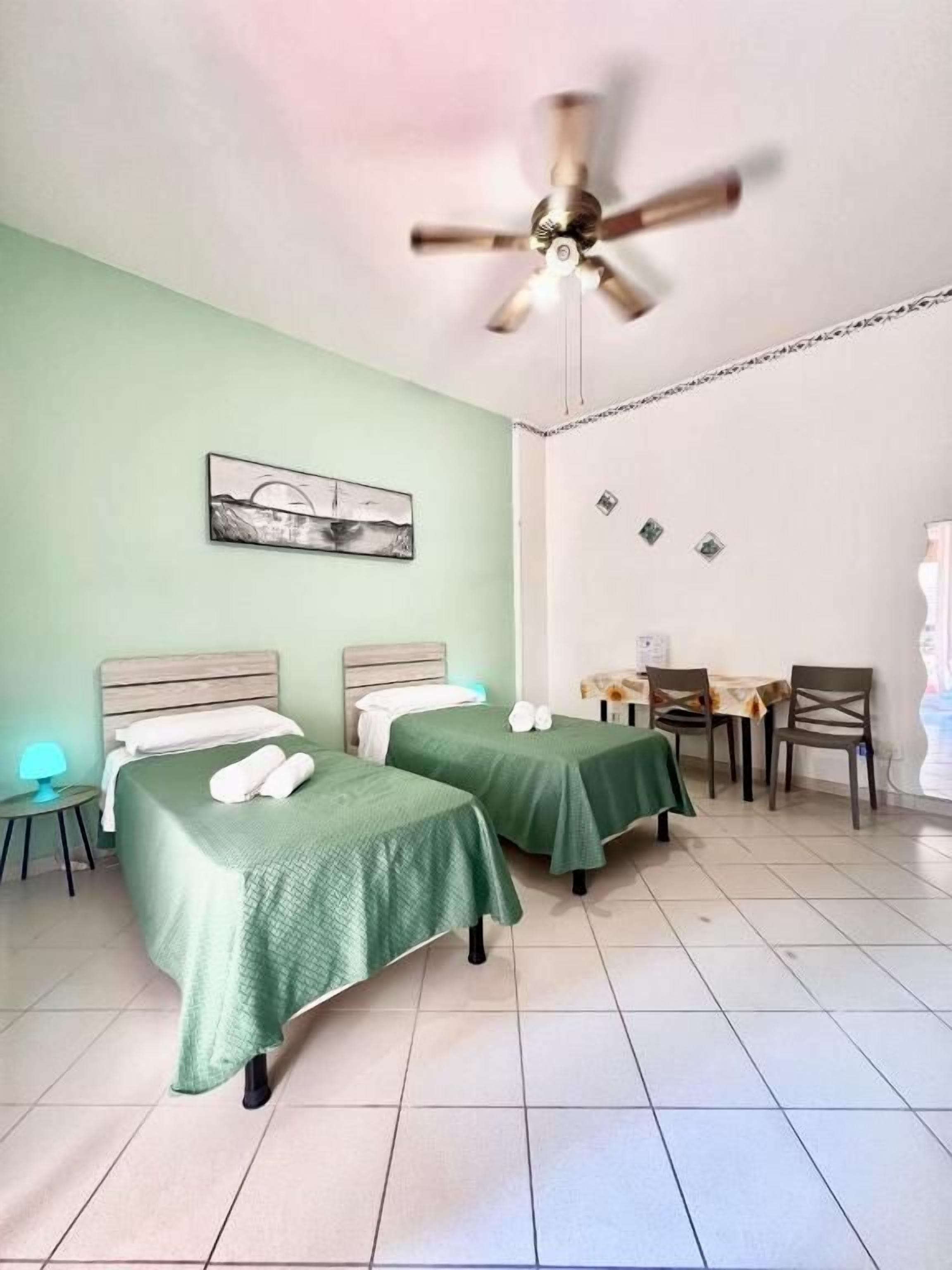 A hotel room with two beds, a ceiling fan, a dining table with chairs, and a small table with a lamp.