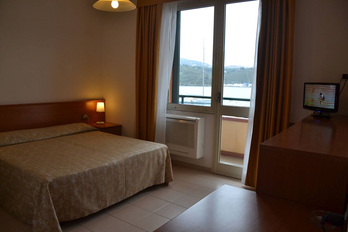 A hotel room with a bed, a desk with a computer, a window with curtains, and a view of a body of water.