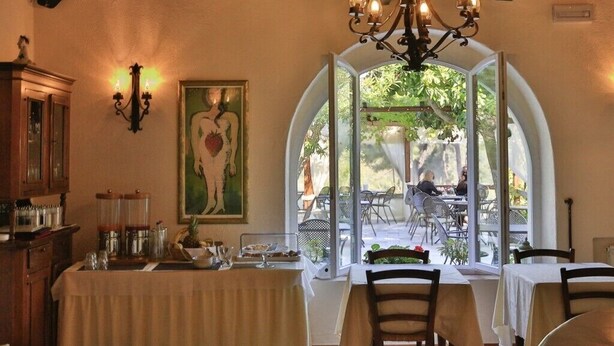 Uma sala de jantar com mesas preparadas para dois, um candelabro, um quadro e vista para o exterior.