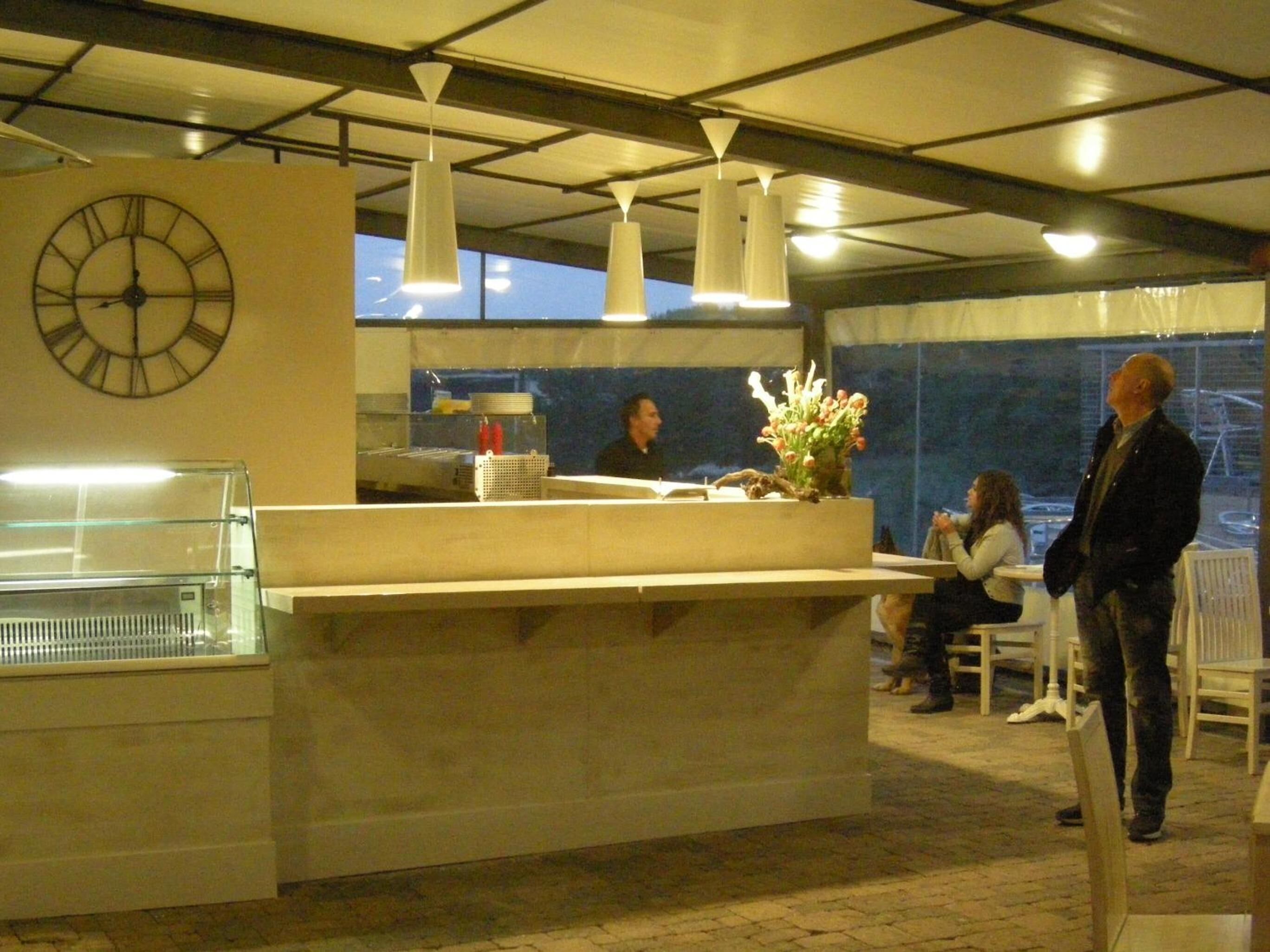 A hotel reception area with a large clock, a counter with a vase of flowers, and a view of the outdoors through large windows.