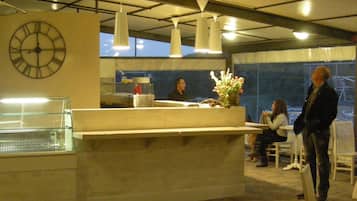 A hotel reception area with a large clock, a counter with a vase of flowers, and a view of the outdoors through large windows.
