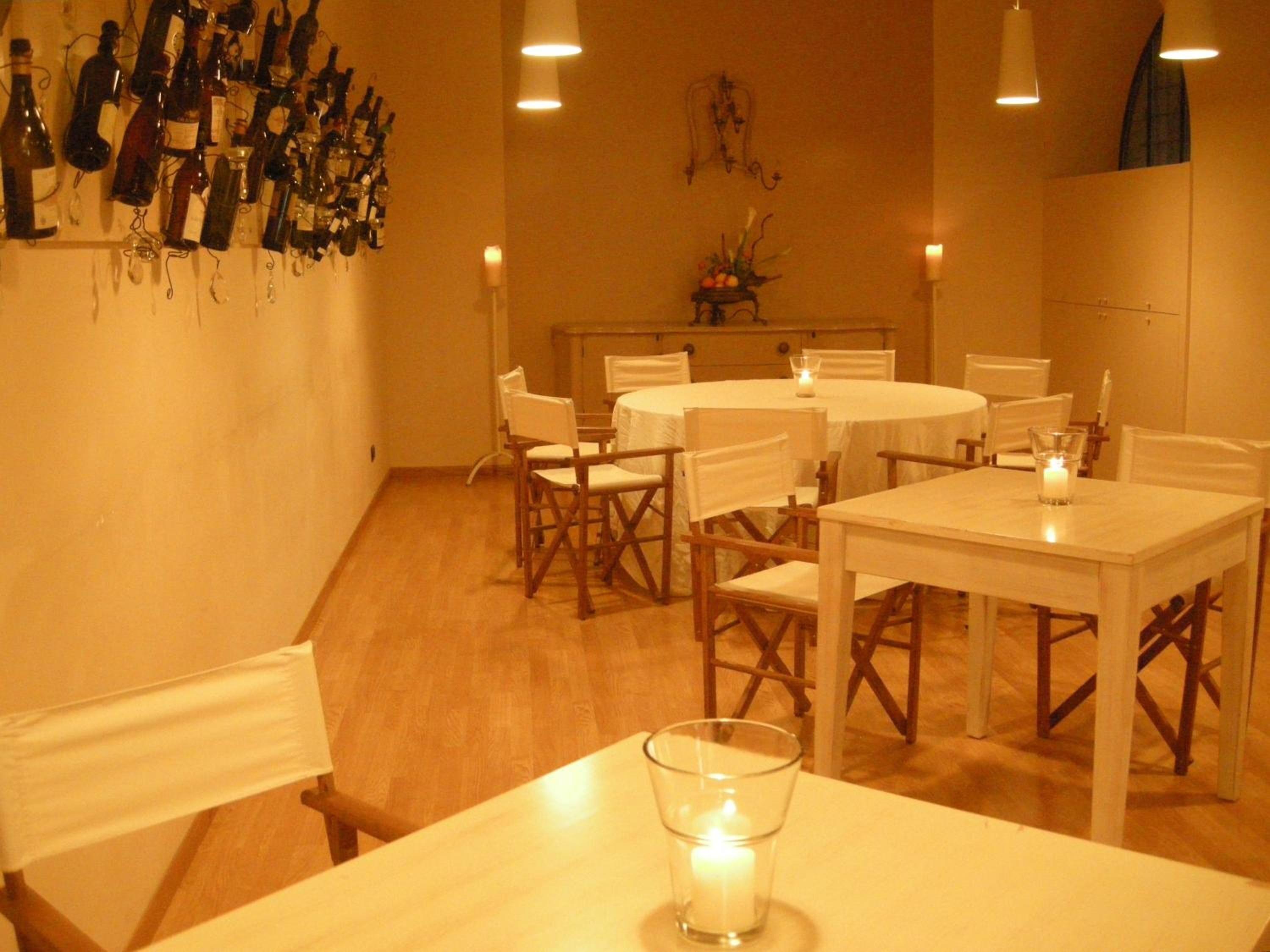 A dining area with white tables and chairs, a wall of wine bottles, and candles on the tables.