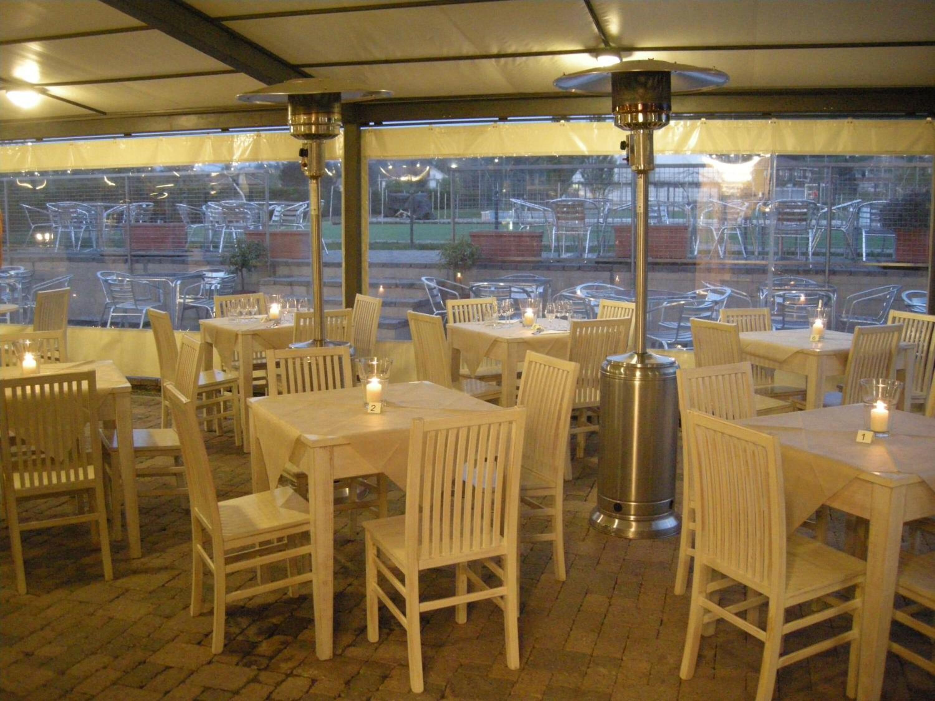 An outdoor dining area with tables and chairs, covered by a canopy, and a view of parked cars outside.