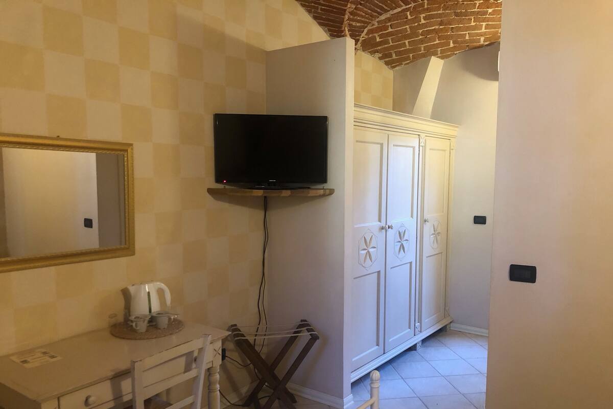 A room with a television mounted on the wall, a white cabinet with decorative doors, a small table with a cup and saucer, and a chair.