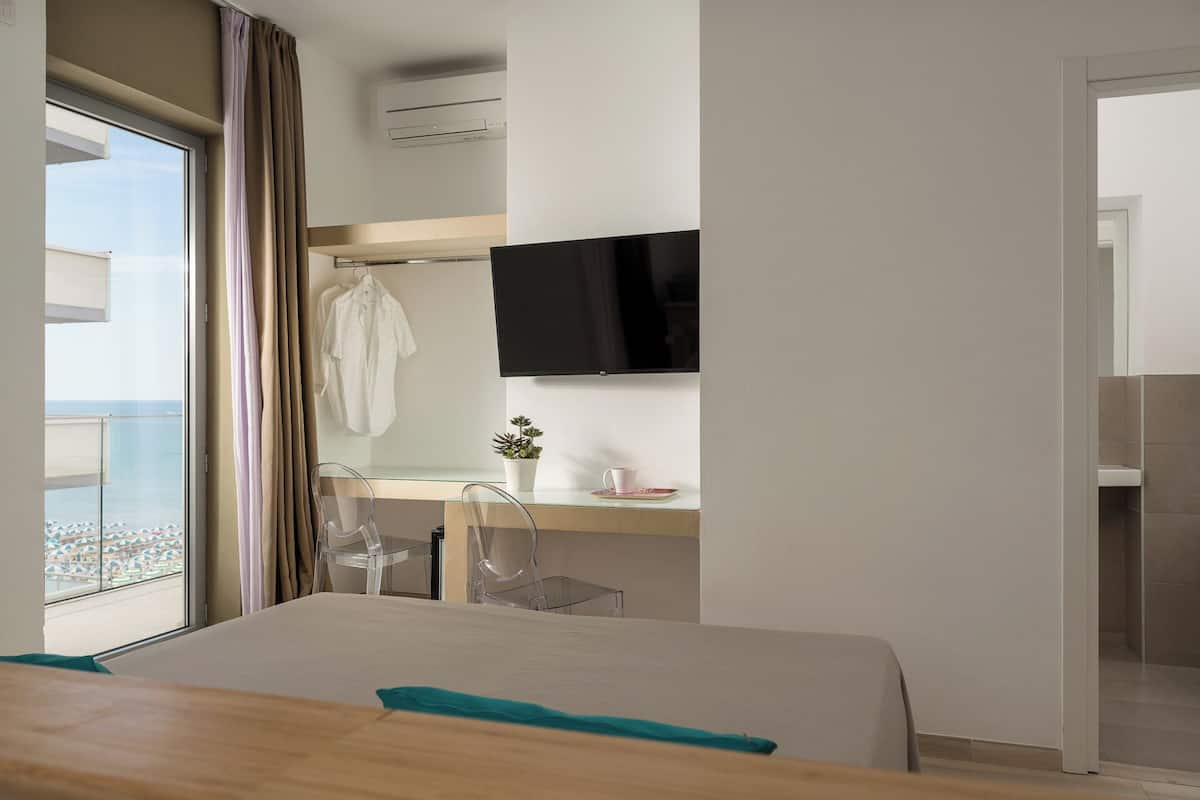 A modern hotel room with a large bed, a desk with a chair, a TV, and a view of the sea.