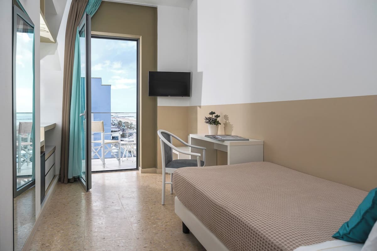 A hotel room with a bed, a desk, a chair, a TV, and a balcony with a view of the city.
