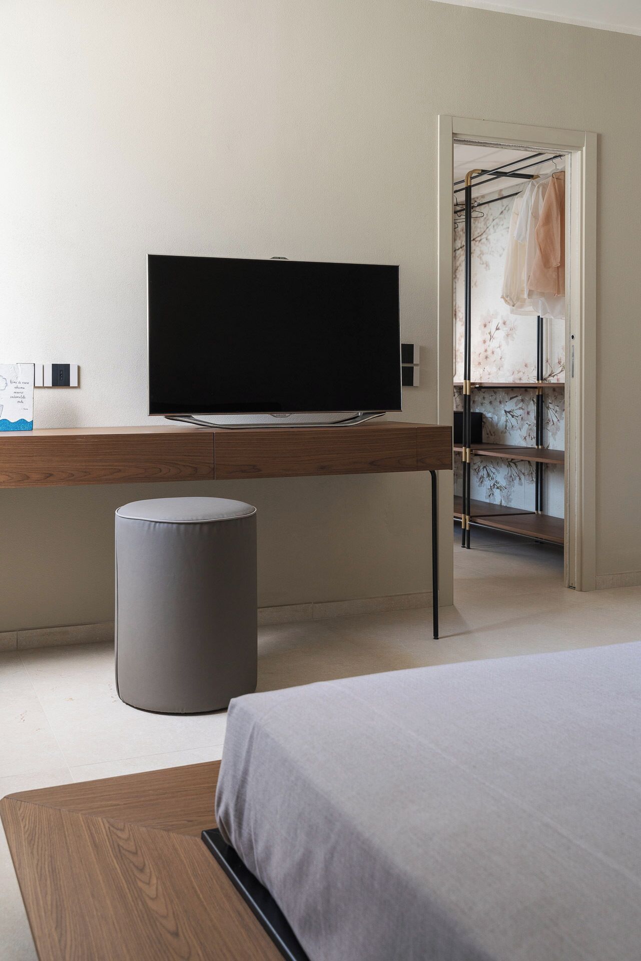 A bedroom with a bed, a desk with a flat-screen TV, a grey trash bin, and a wardrobe with a floral design.
