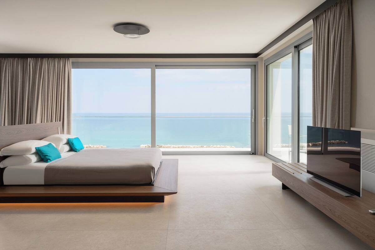 A modern bedroom with a large bed, a TV, and a view of the ocean.